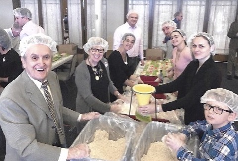 St. Mary's Parishioners stand around a meal packing station table smiling, ready to package oatmeal for Harvest Pack.